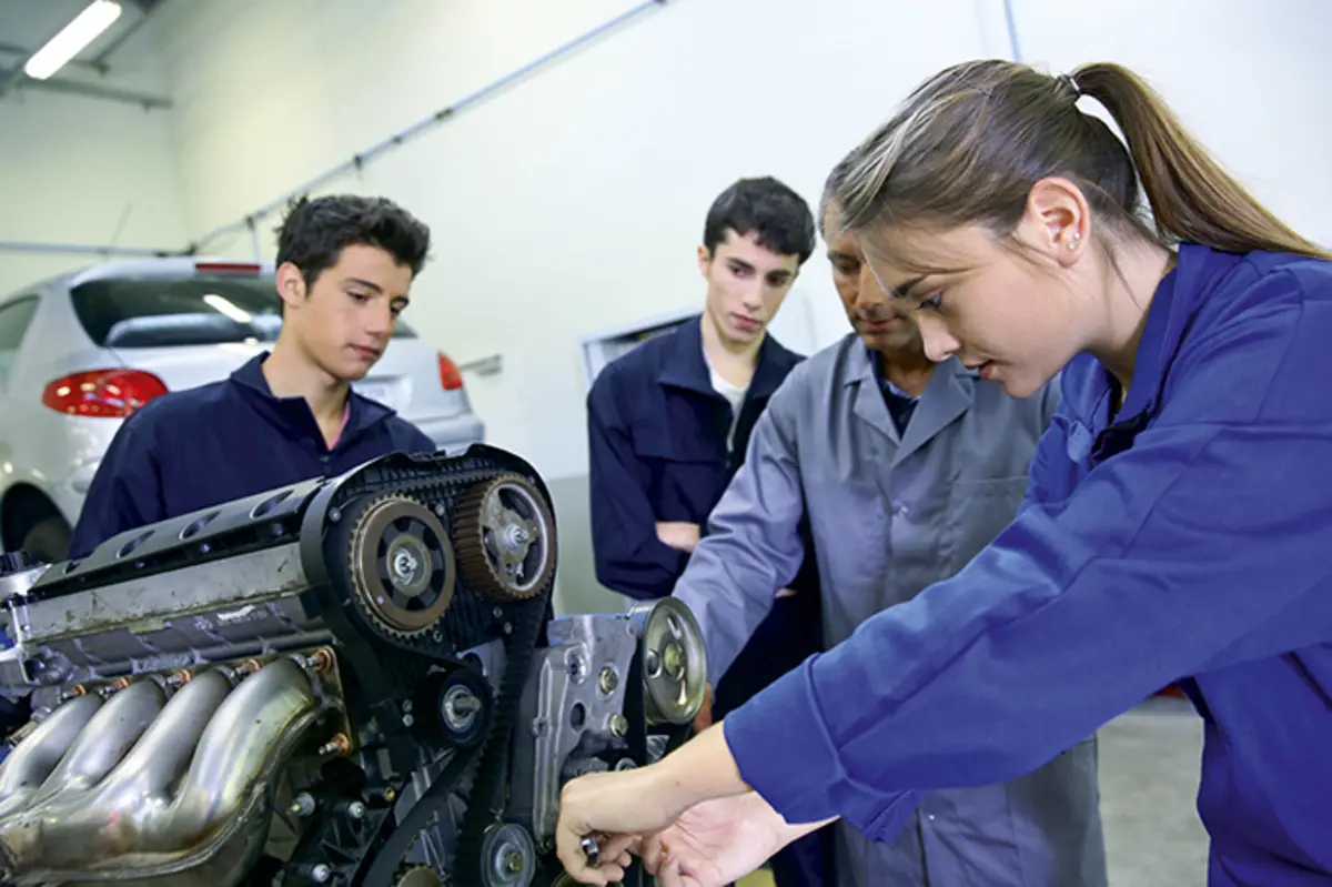 2 jeunes hommes et une jeune femme autour d'un professeur et d'un moteur de voiture. La jeune fille utilise un ouils, les jeunes hommes regardent