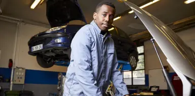 Lukau pose devant un capot ouvert. En arri&egrave;re plan une voiture mont&eacute; sur v&eacute;rin