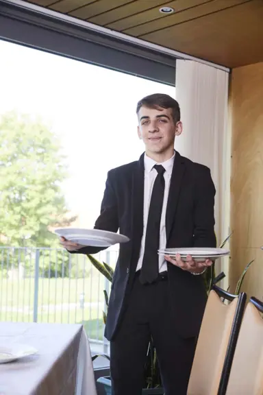 Maxence, en costume cravatte, porte une assiette dans chaque main