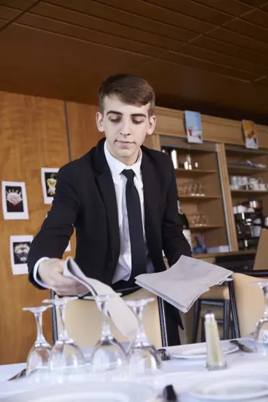 Maxence, en costume cravatte, pose des serviettes en tissu sur une table dress&eacute;e d'assiettes et de verres &agrave; pied