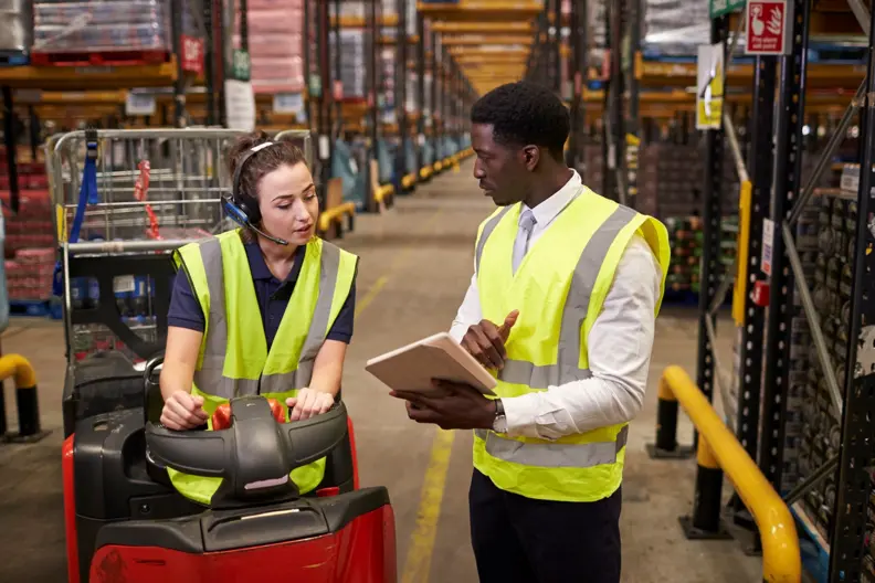 Une femme assise sur un engin de logistique. 1 homme &agrave; ses c&ocirc;t&eacute; tient un carnet &agrave; la main. Ils discutent