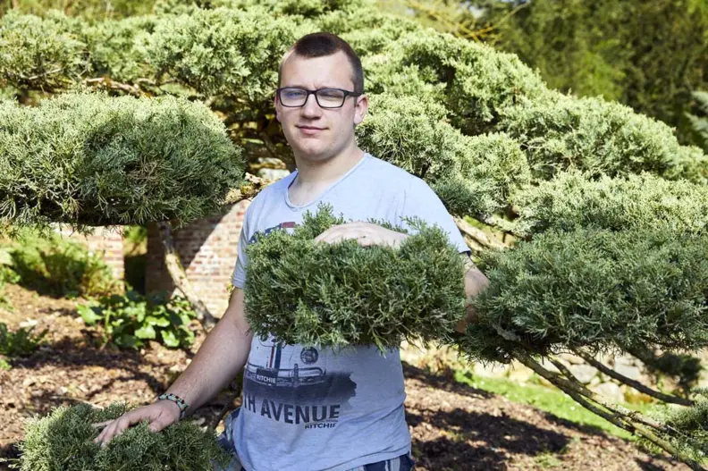 Un jeune homme pose devant un buisson. Il a sa main dessus