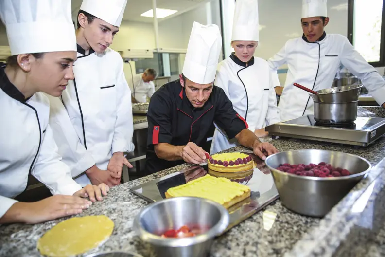 Un cuisinier d&eacute;pose des framboises sur un g&acirc;teau rond. 4 personnes autour de lui le regardent